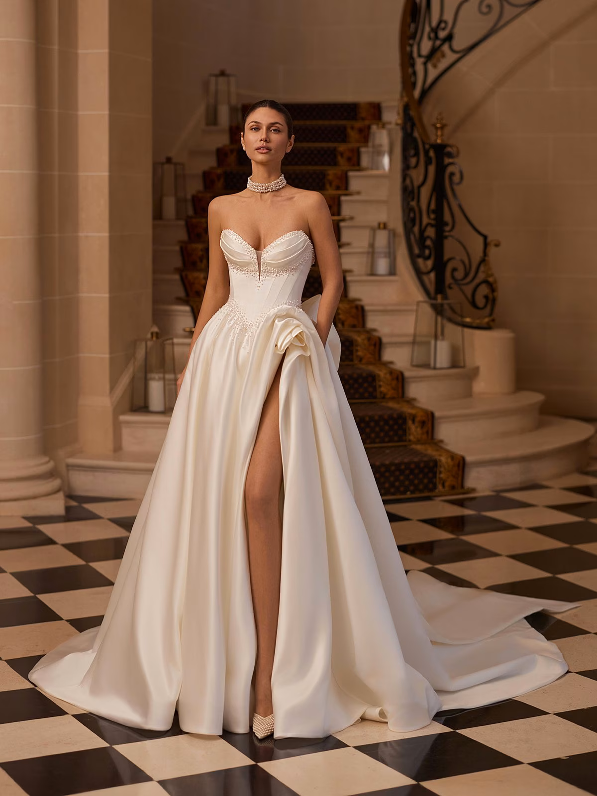 Val Stefani ASTRAELIA Elegant bride in a strapless ivory wedding gown with a high slit, scattered pearls along pointed sweetheart A-line wedding dress standing on a black and white checkered floor and a grand staircase in the background