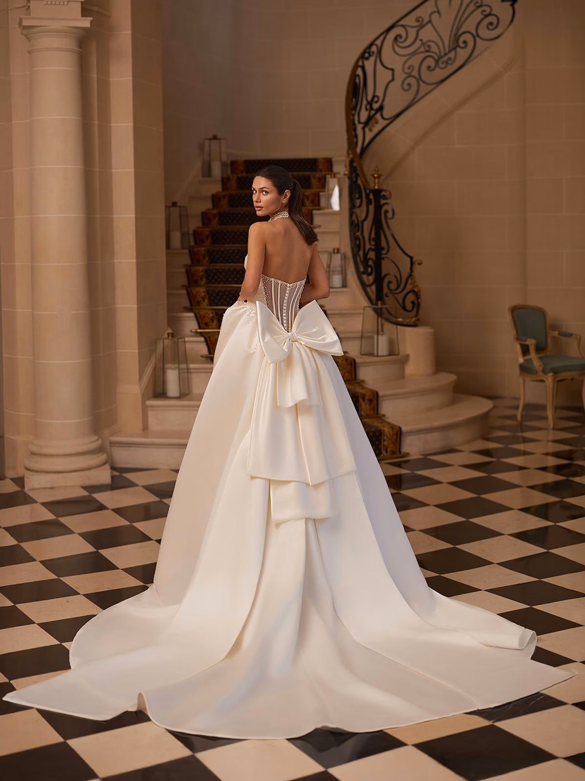 A bride in an elegant strapless white gown with a large bow stands on black-and-white checkered floor, with a grand staircase in the background Val Stefani ASTRAELIA 