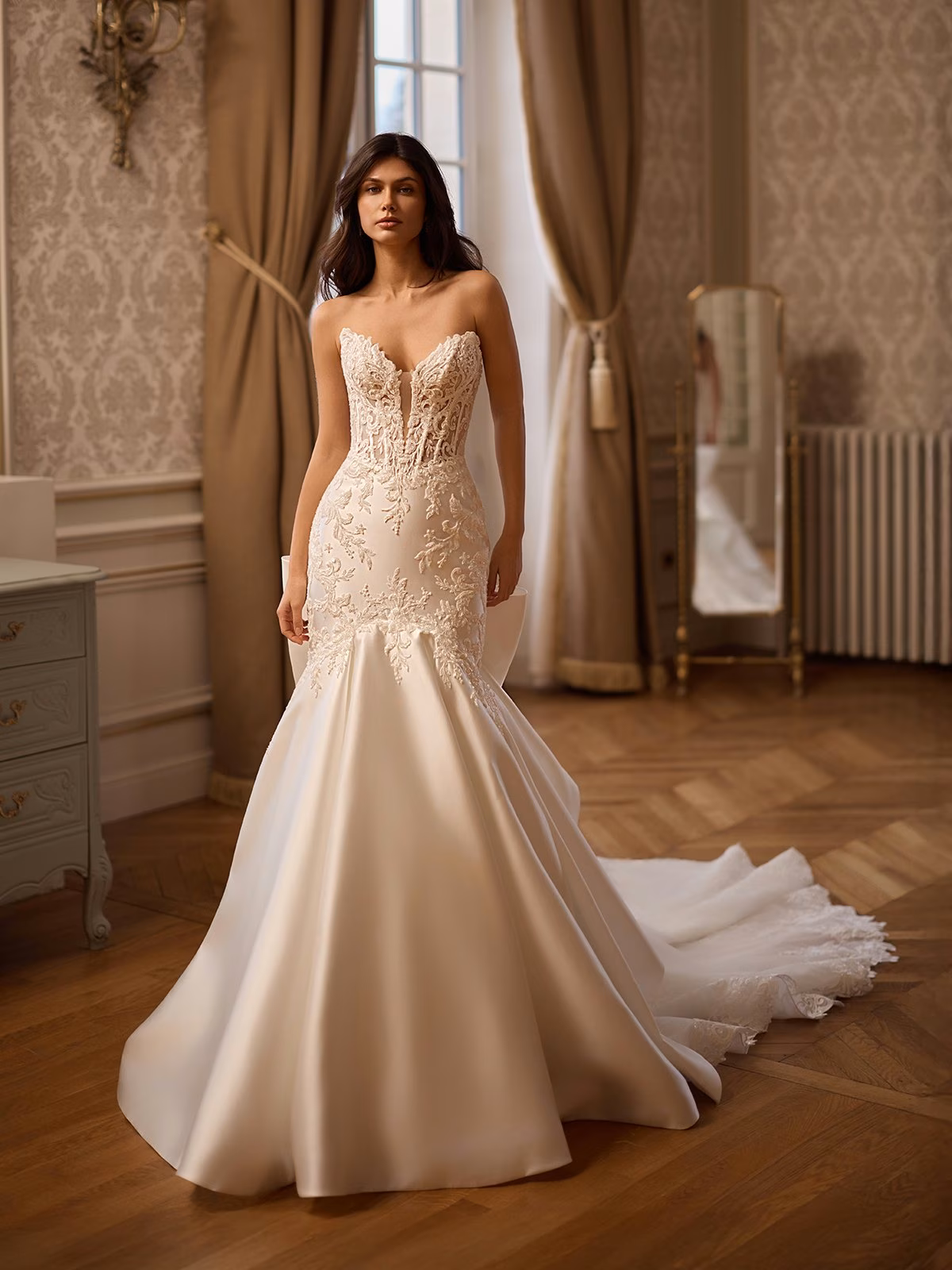 A bride stands elegantly in a vintage room, wearing a strapless, lace-embellished ivory mermaid wedding gown. The setting is warm with ornate wallpaper and wooden floors Val Stefani CAMBRIDGE 
