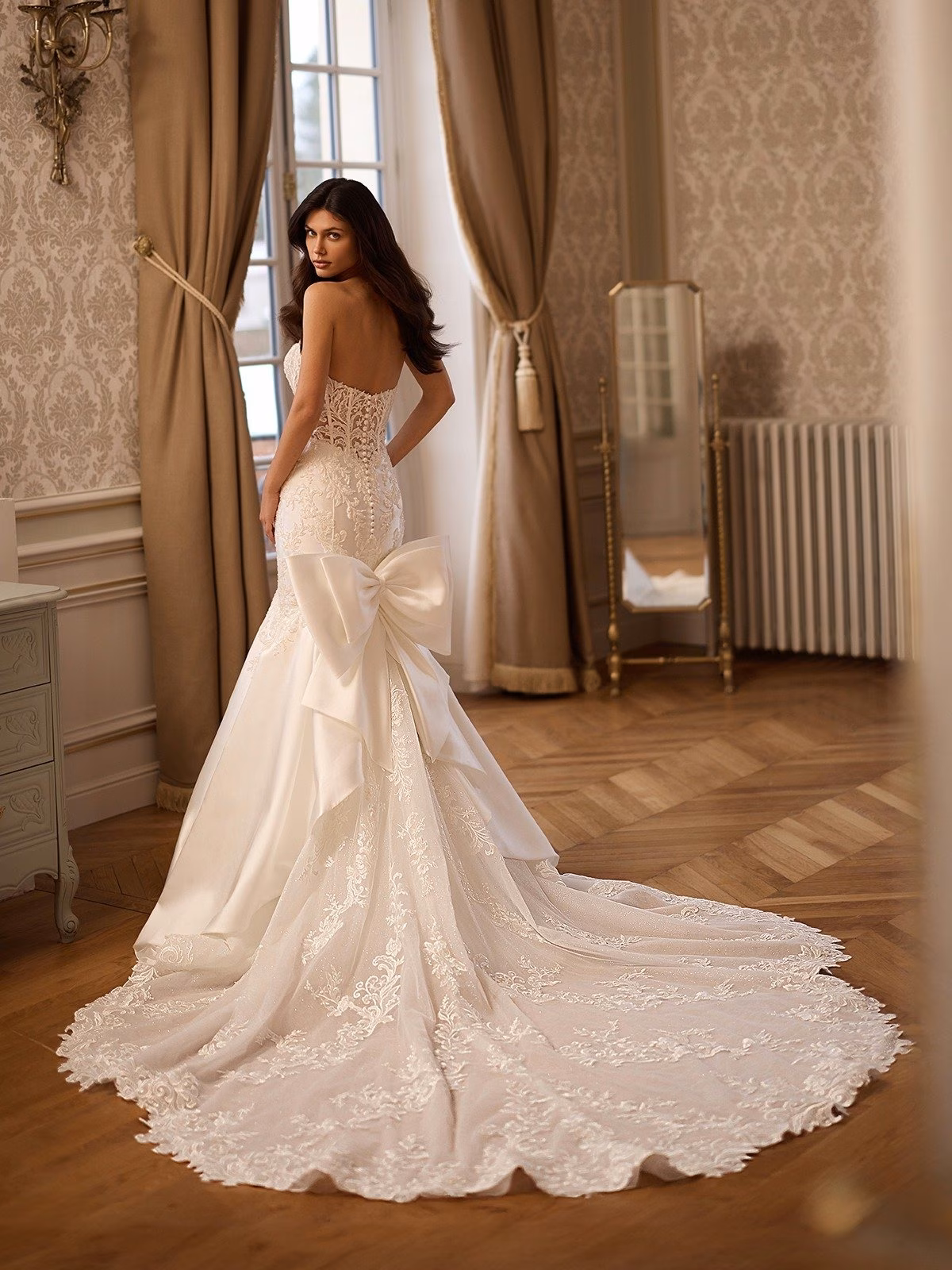 A woman in an elegant, strapless bridal gown with lace details stands in a vintage room. The dress has a long train and large bow, exuding timeless grace Val Stefani CAMBRIDGE