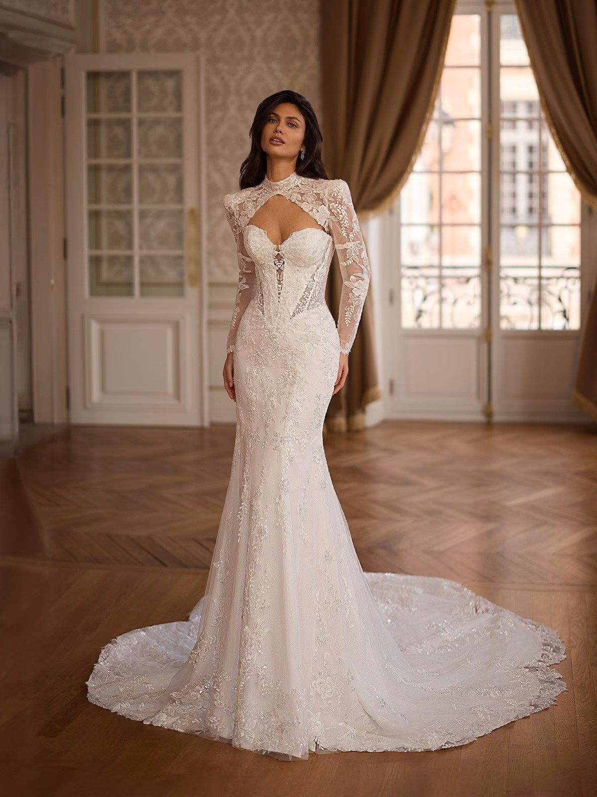 Bride in elegant mermaid lace wedding gown with bolero jacket stands in a classy room with parquet flooring and large windows. The dress exudes sophistication and grace Val Stefan Duchess