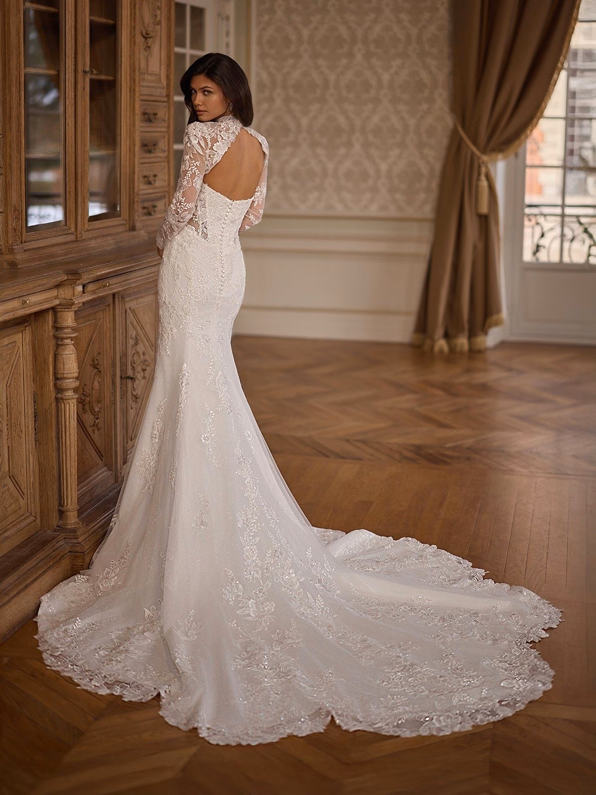 Bridal gown with lace detailing, long sleeves, and open back, flowing lace Chapel train on hardwood floor. Elegant setting with vintage wooden cabinet and draped window Val Stefani DUCHESS