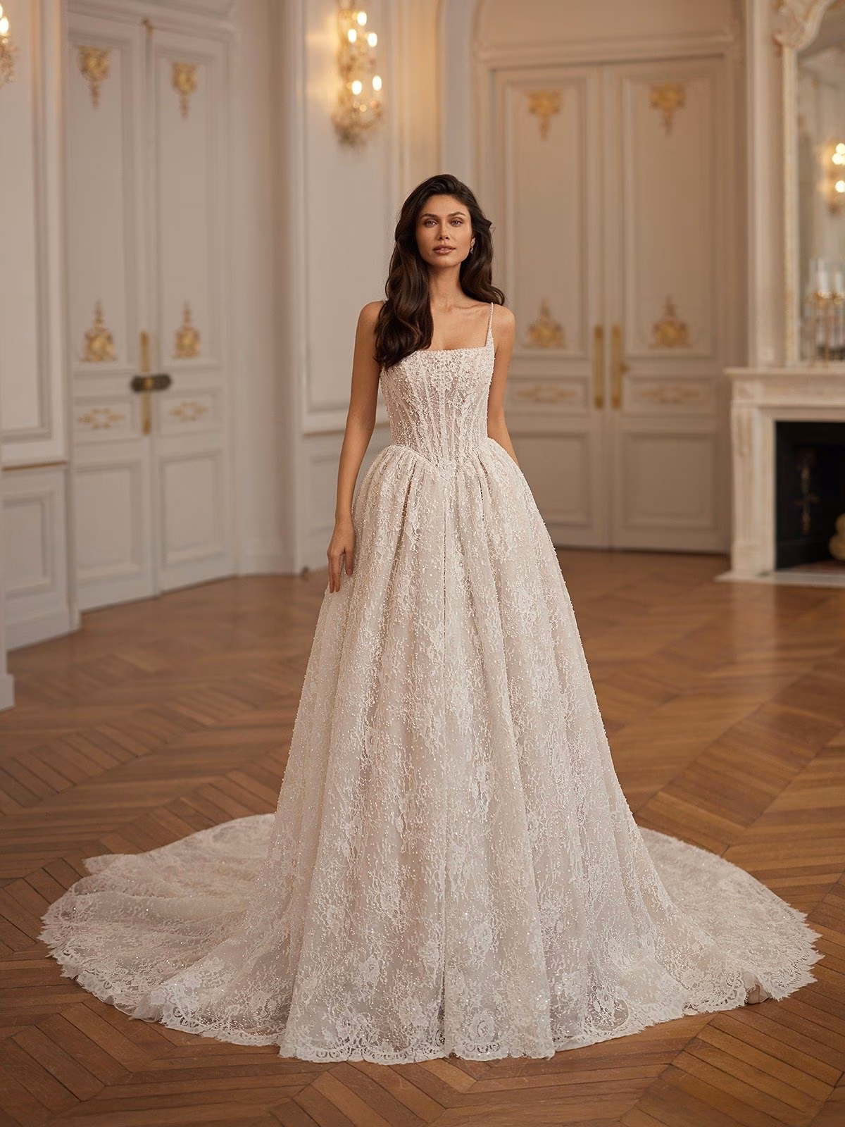 A bride stands in an elegant room wearing a square neck beaded lace wedding gown with a Basque waist. The room features ornate white doors and warm lighting Val Stefani HIGHGROVE