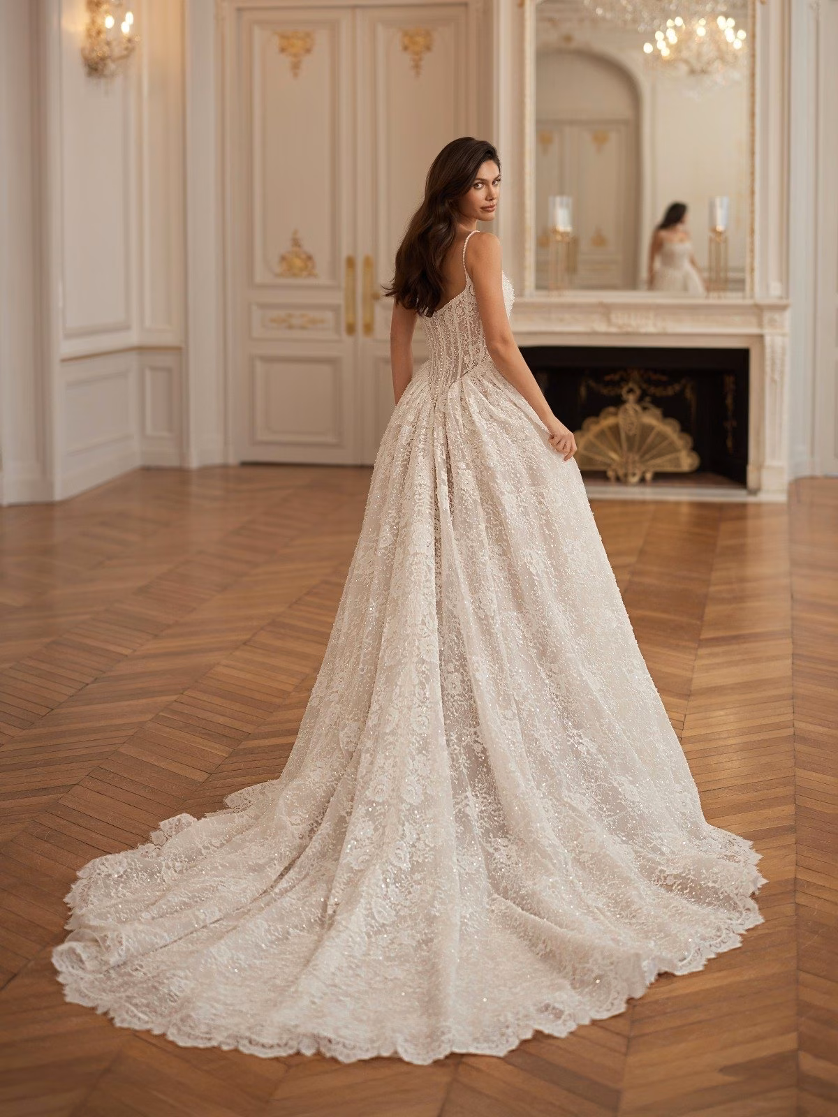 A bride in a flowing beaded lace long train wedding gown stands elegantly in a luxurious room with ornate details, wood flooring, and a large mirror. The atmosphere is sophisticated.Val Stefani HIGHGROVE