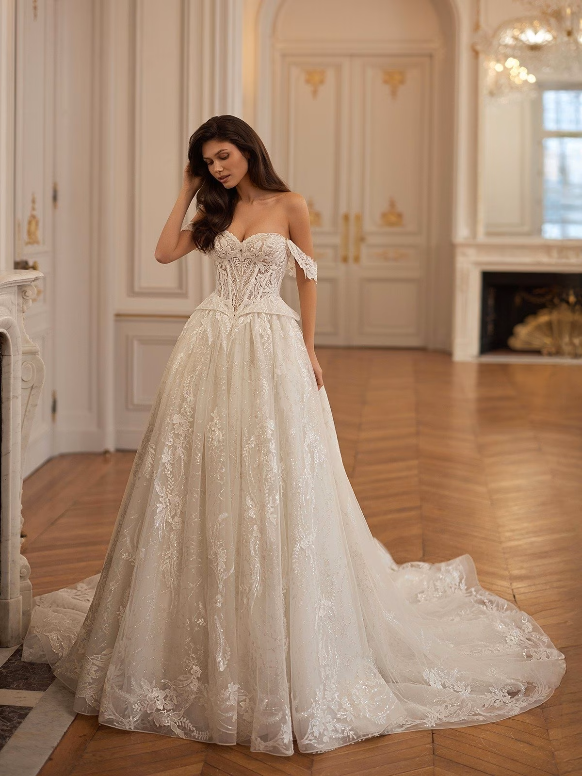 A bride in an opulent off-shoulder lace sparkly wedding gown stands in a luxurious room with wooden floors and chandeliers, exuding romance and grace Val Stefani LUXANA