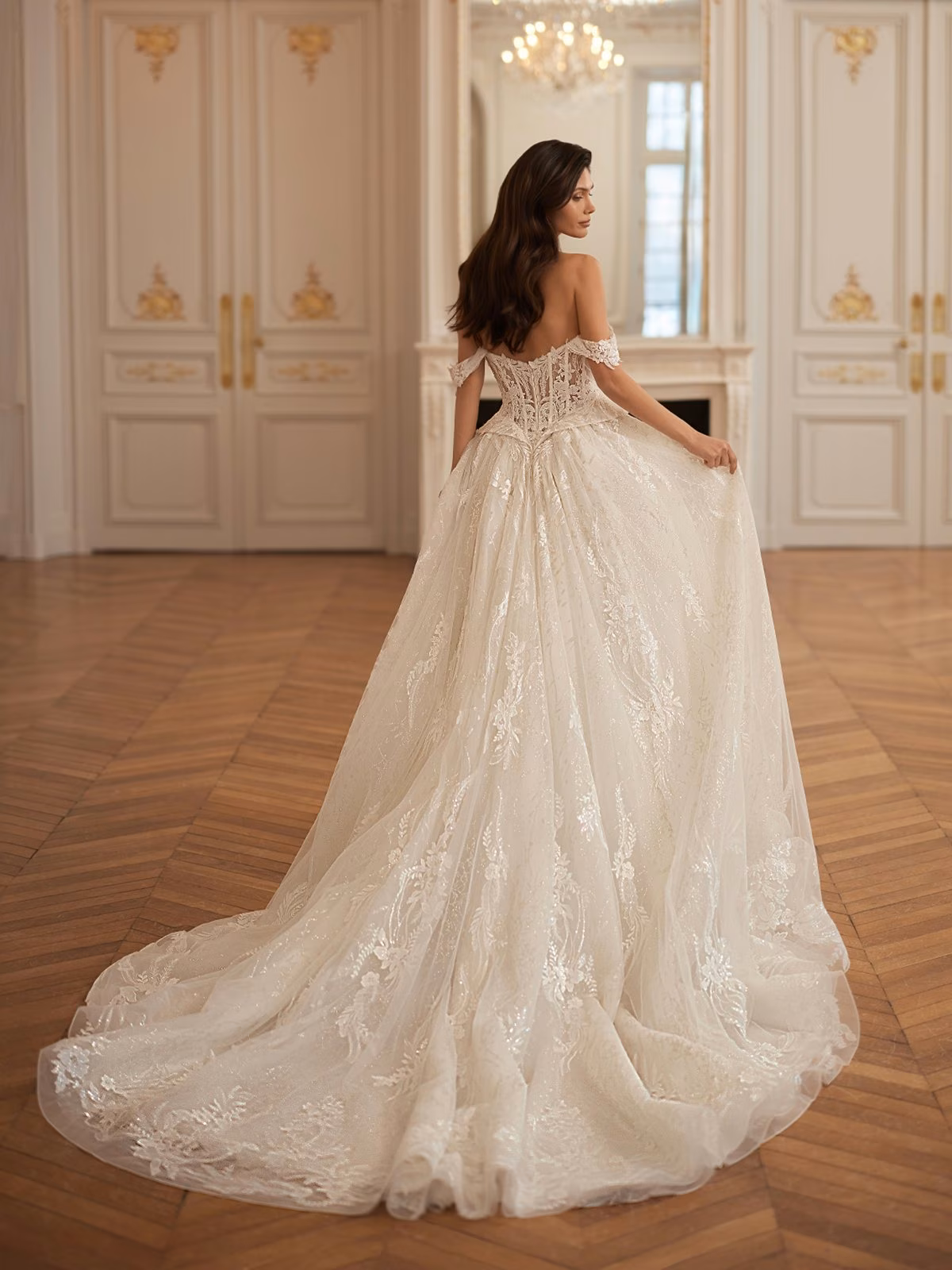 Bride in an elegant off-the-shoulder lace and sparkly gown stands in an ornate ballroom, walking away. The room has intricate white walls and a wooden floor. Val Stefani LUXANA 