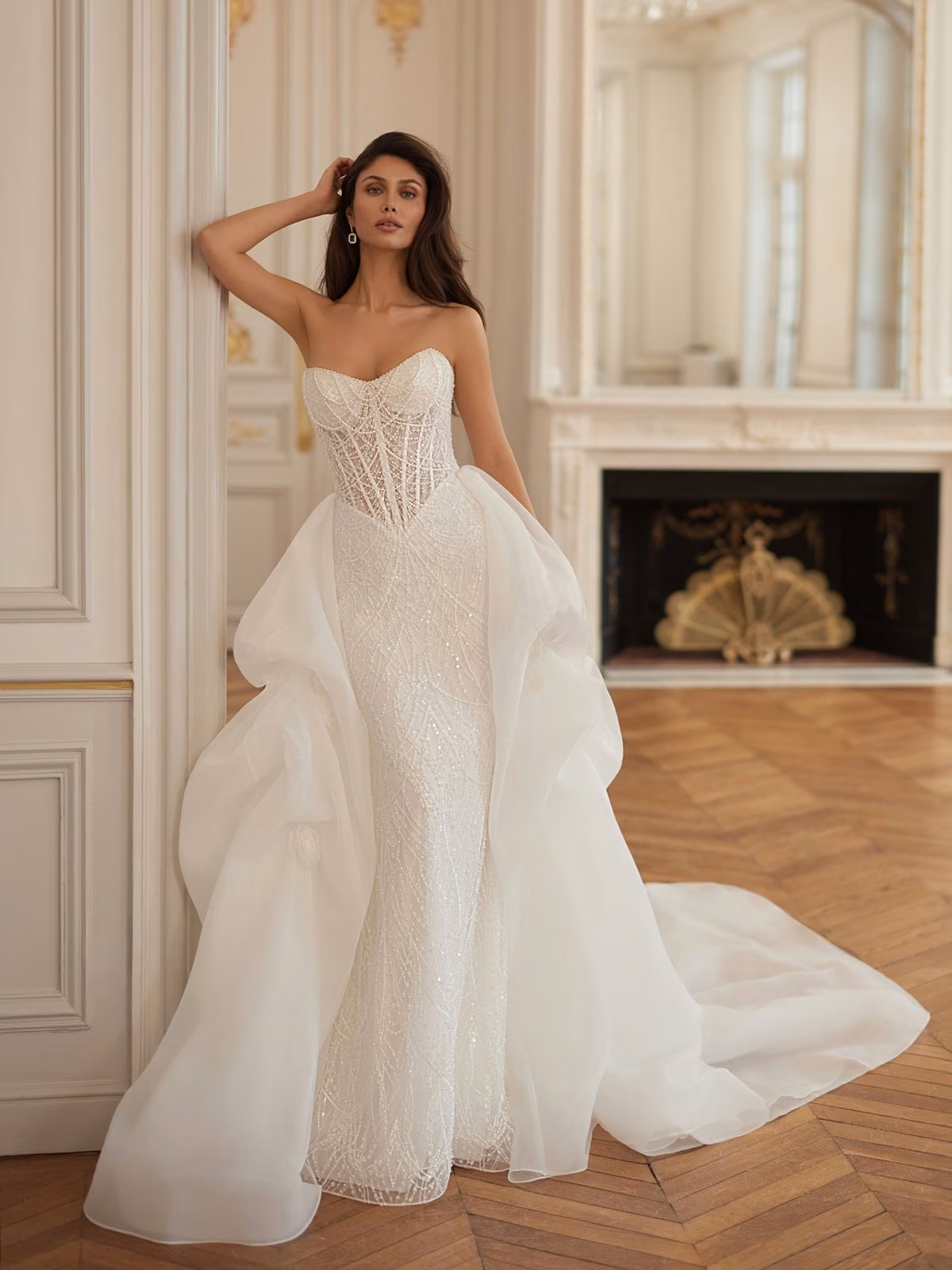 Elegant bride in a strapless, intricately beaded wedding gown with flowing detachable train, leaning against a classic ornate wall in a luxurious, airy room Val Stefani REGALIA 